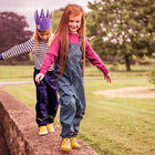 Two young girls balancing on a wall in a park. The girl in front wears green Originals Waterproof Dungarees and yellow boots.